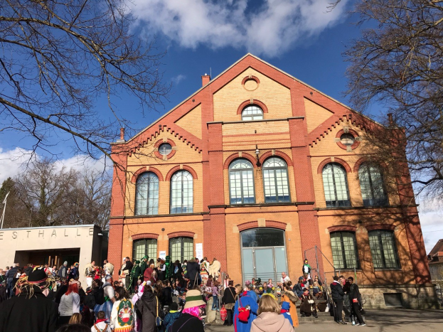 Eine große Gruppe von Menschen, die vor einem mehrstöckigen Gebäude mit Fenstern und Türen im Freien stehen, umgeben von Bäumen unter einem bewölkten Himmel, möglicherweise an einem Festival teilnehmen.