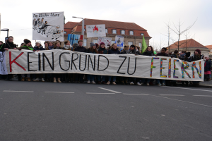Demonstranten halten ein Transparent mit der Aufschrift 'Kein Grund zur Freude' gegen deutsche Sparmaßnahmen, mit Straßeninfrastruktur und Gebäuden im Hintergrund.