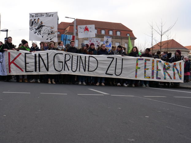 Demonstranten halten ein Transparent mit der Aufschrift 'Kein Grund zur Freude' gegen deutsche Sparmaßnahmen, mit Straßeninfrastruktur und Gebäuden im Hintergrund.