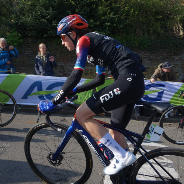 Die professionelle Radrennfahrerin Katie Archibald fährt ein Fahrrad auf einer Straße, trägt einen Helm, eine Schutzbrille, Handschuhe und Schuhe und fährt an einer Zuschauermenge vorbei.