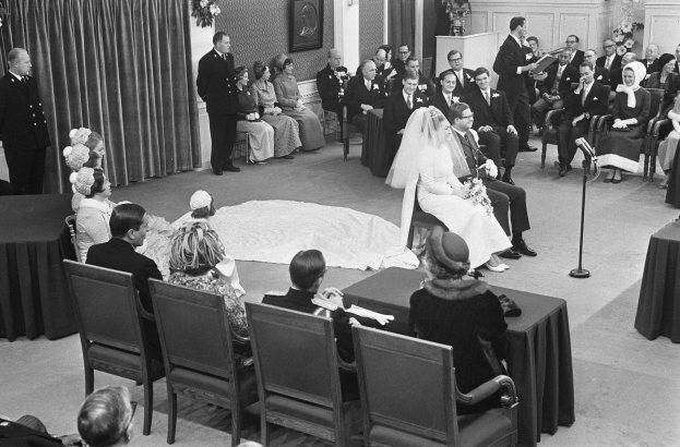 Schwarzes und weißes Foto einer Hochzeitszeremonie mit Menschen, die sitzen und stehen, einem Mann mit Mikrofon, einer Frau mit einem Strauß Blumen, einem Podium mit Mikrofon, Blumenschmuck, einem Vorhang, einem Rahmen an der Wand und einer Tür im Hintergrund.