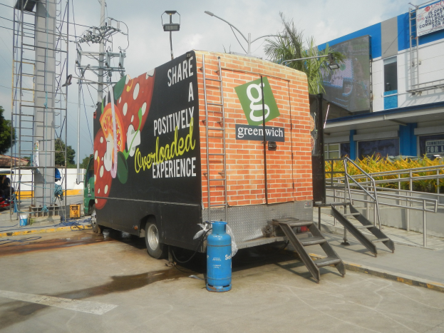 Lastwagen vor einem Gebäude mit einer Tafel, auf der "Teilen Sie eine positiv überladene Erfahrung" steht, mit Stufen, einem Zylinder auf dem Boden, Geländern, Strommasten mit Drähten, Laternenmasten, Bäumen und einem bewölkten Himmel im Hintergrund.