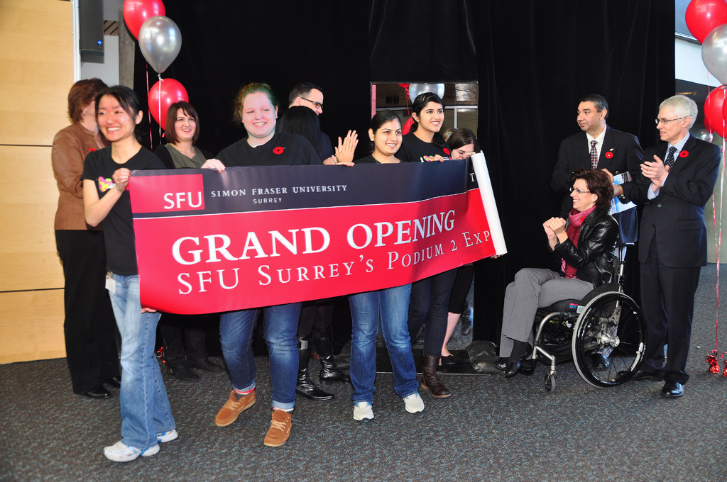 Eine Gruppe von Frauen hält ein Banner, auf dem ein Event beworben wird, daneben stehen zwei klatschende Männer und eine aufgeregte Frau im Rollstuhl vor einem schwarzen Vorhang und Ballons.