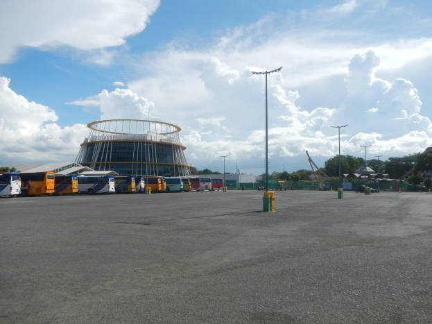 Ein Parkplatz mit zahlreichen Bussen vor einem Gebäude, umgeben von Straßenmasten, Straßenlaternen, Bäumen und einem bewölktem Himmel.
