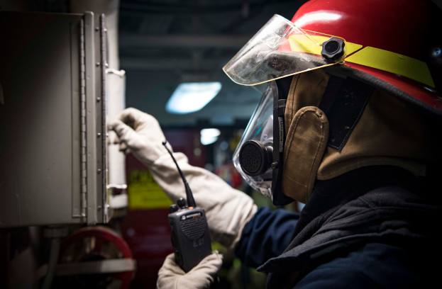 Ein Feuerwehrmann in voller Montur hält ein Walkie-Talkie und ein Metallobjekt, mit einem unscharfen Hintergrund.