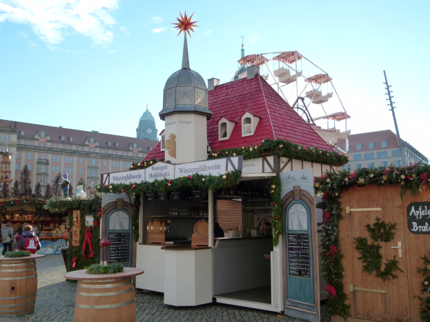 Kleines Gebäude mit rotem Dach und Riesenrad davor, umgeben von festlichen Oktoberfest-Dekorationen, Menschen und Gegenständen, vor einer Kulisse aus Gebäuden, Bäumen und bewölktem Himmel.