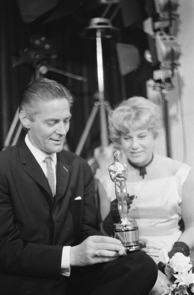 Ein Mann in einem schwarzen Blazer, weißem Hemd und Krawatte hält eine Oscar-Statue in der Hand und sitzt neben einer Frau, die einen Blumenstrauß hält, beide in eleganter Kleidung, mit ein paar Lichtern im Hintergrund, dargestellt in Schwarz-Weiß.