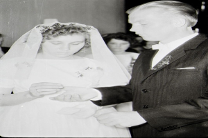 Ein Schwarz-Weiß-Foto einer Braut in einem weißen Kleid und Schleier und eines Bräutigams in einem Anzug, die Ringe tauschen, mit ein paar Leuten im Hintergrund.