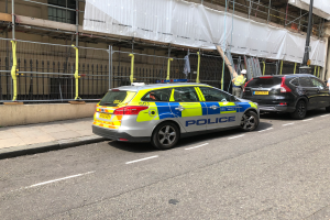 Ein Polizeiwagen steht an der Seite einer Straße neben einem Gebäude, daneben stehen zwei Helme tragende Personen; im Vordergrund sind ein Geländer und ein Pfahl zu sehen, im Hintergrund ein weißes Tuch über das Gebäude drapiert, was auf eine polizeiliche Untersuchung nach einem Messerangriff in London hindeutet.