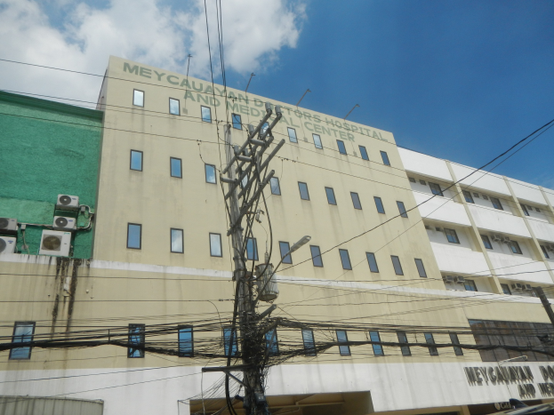 Großes Gebäude mit vielen Fenstern, ein elektrischer Mast mit zahlreichen Drähten und ein bewölkter Himmel im Hintergrund; das Gebäude zeigt eine Beschriftung, die es als Krankenhaus identifiziert.