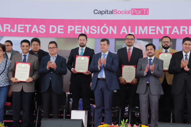 Eine Gruppe von Menschen auf einer Bühne, die klatschen und Zertifikate halten, mit Stühlen in einem Halbkreis und einem Banner mit der Aufschrift "Capital Social for Ti" im Hintergrund sowie Blumensträuße und Redner unten.