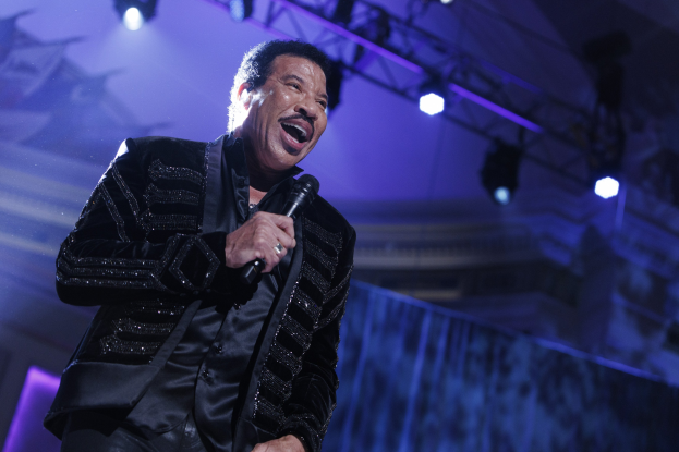 Lionel Richie performs bei den Grammys, tr├Ągt ein schwarzes Kleid und h├Ąlt ein Mikrofon in der Hand, mit hellen Scheinwerfern, die eine dramatische Atmosph├Ąre schaffen.
