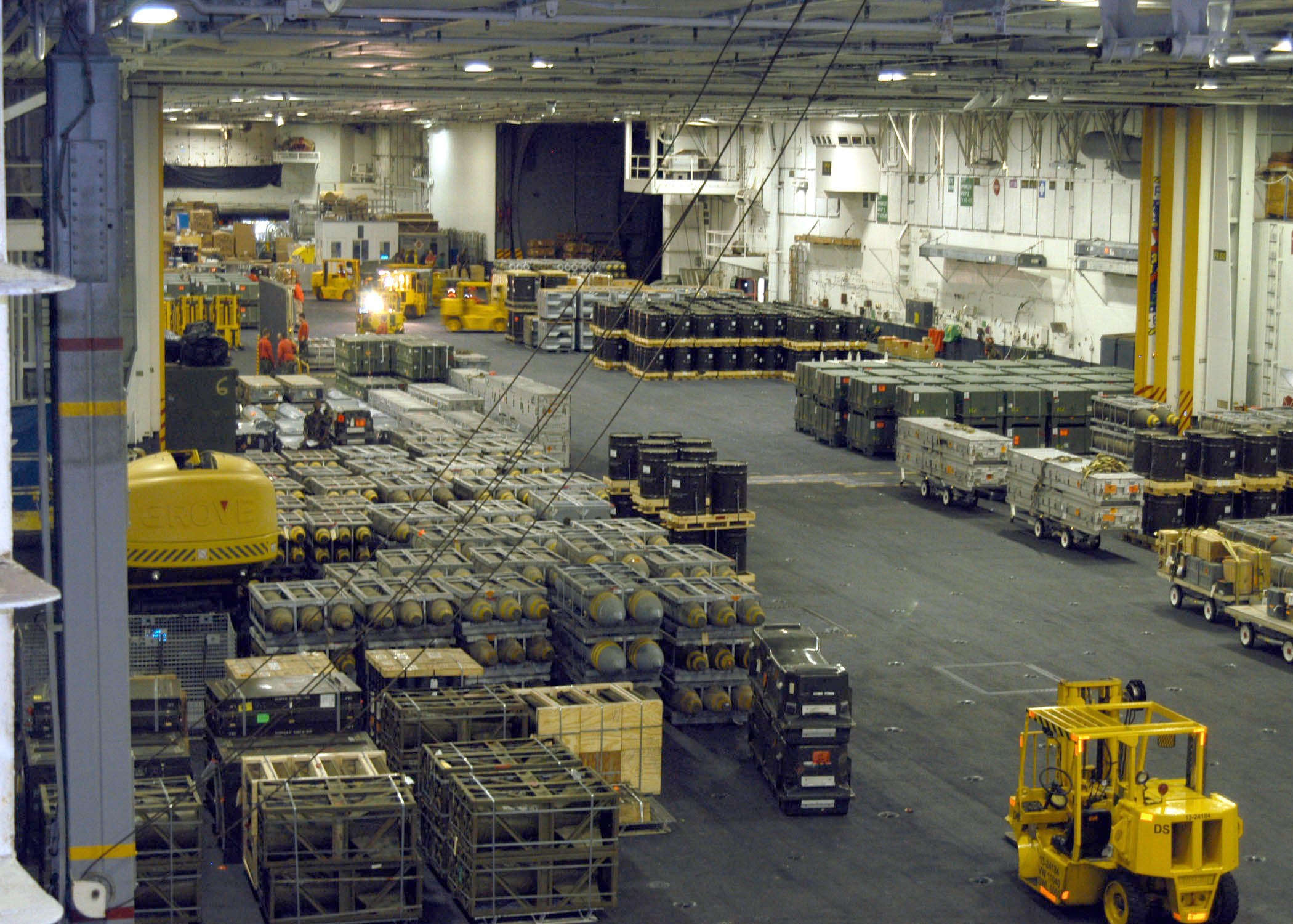 Großes Lagerinnere mit mehreren Lastwagen, Containern, Menschen, Deckenbeleuchtung, Metallstangen und einem sichtbaren Rad.