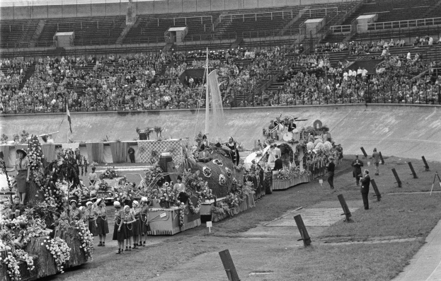 Schwarzes und weißes Foto eines Umzugs in einem Stadion mit Menschen, die stehen und sitzen, einem zentralen Springbrunnen und Blumengebinden auf Fahrzeugen.