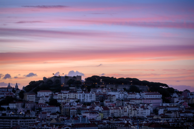 Sonnenuntergangsblick auf Lissabon, Portugal von einem Hügel aus, mit Gebäuden und Bäumen im Vordergrund und Wolken am Himmel.