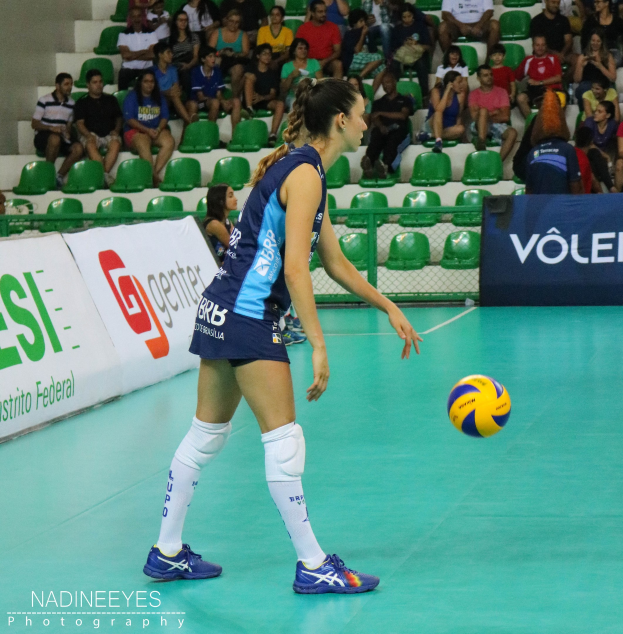 Frauen-Volleyballspielerin in Aktion auf einem Court mit einem Ball in der Luft, vor dem Hintergrund sitzender Zuschauer, Werbetafeln, Stufen und einer Wand.