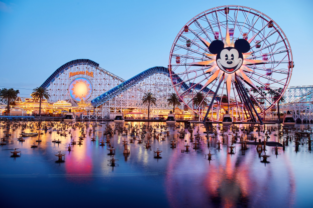 Eine farbenfrohe Dämmerungsszene im Disneyland California Adventure mit einem riesigen Mickey-Maus-Rad im Vordergrund, einer Achterbahn im Hintergrund und Booten auf dem Wasser, mit einem farbenfrohen Himmel und silhouettenartigen Bäumen.
