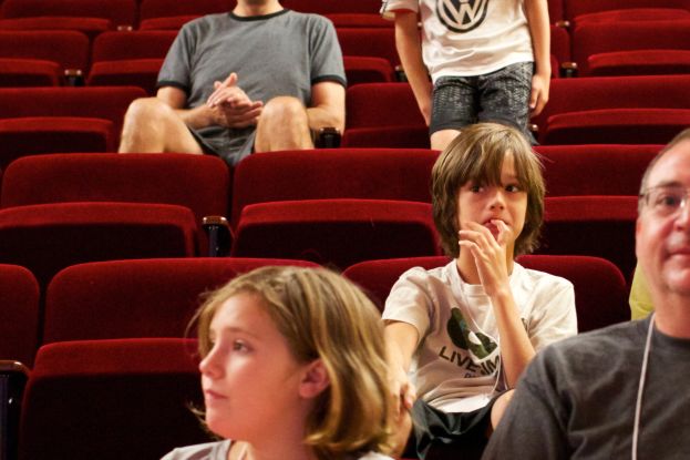 Eine Gruppe von Menschen in einem Kinosaal mit roten Stühlen, ein Junge in einem weißen T-Shirt und schwarzen Hosen in der Mitte.