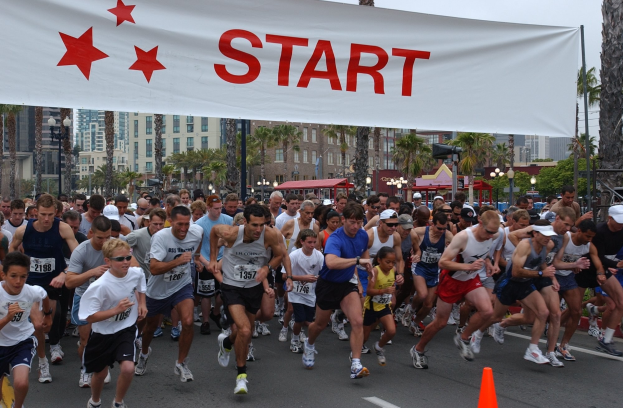 Gruppe von Läufern bei einem Marathon mit einem Verkehrskegel im Vordergrund und einer Fahne mit Text im Hintergrund, umgeben von Bäumen, Laternenmasten, Gebäuden und einem klaren blauen Himmel.