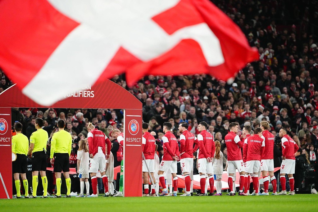 Gruppe von Menschen auf einem Fußballfeld mit einer roten und weißen Flagge im Vordergrund, einem Bogen mit der Aufschrift "Bayern München vs. Bayern München Wetten Tipps & Vorschau" im Hintergrund und einer großen Menschenmenge im Stadion.