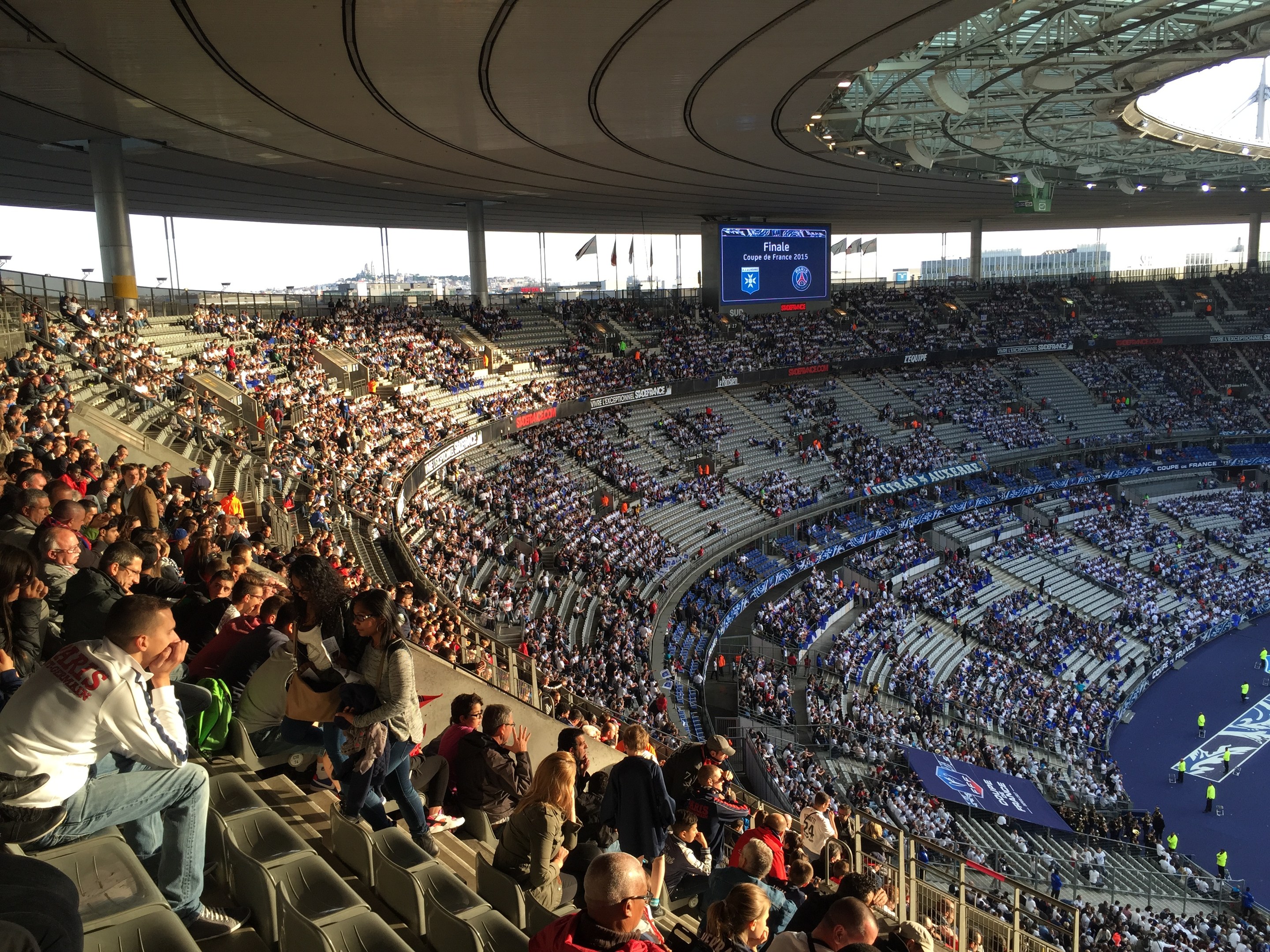 Große Zuschauermenge in einem Stadion bei einem Fußballspiel, mit einer Bühne rechts, Fahnen, Stangen, einem Bildschirm und der Allianz Arena in München, Deutschland im Hintergrund.